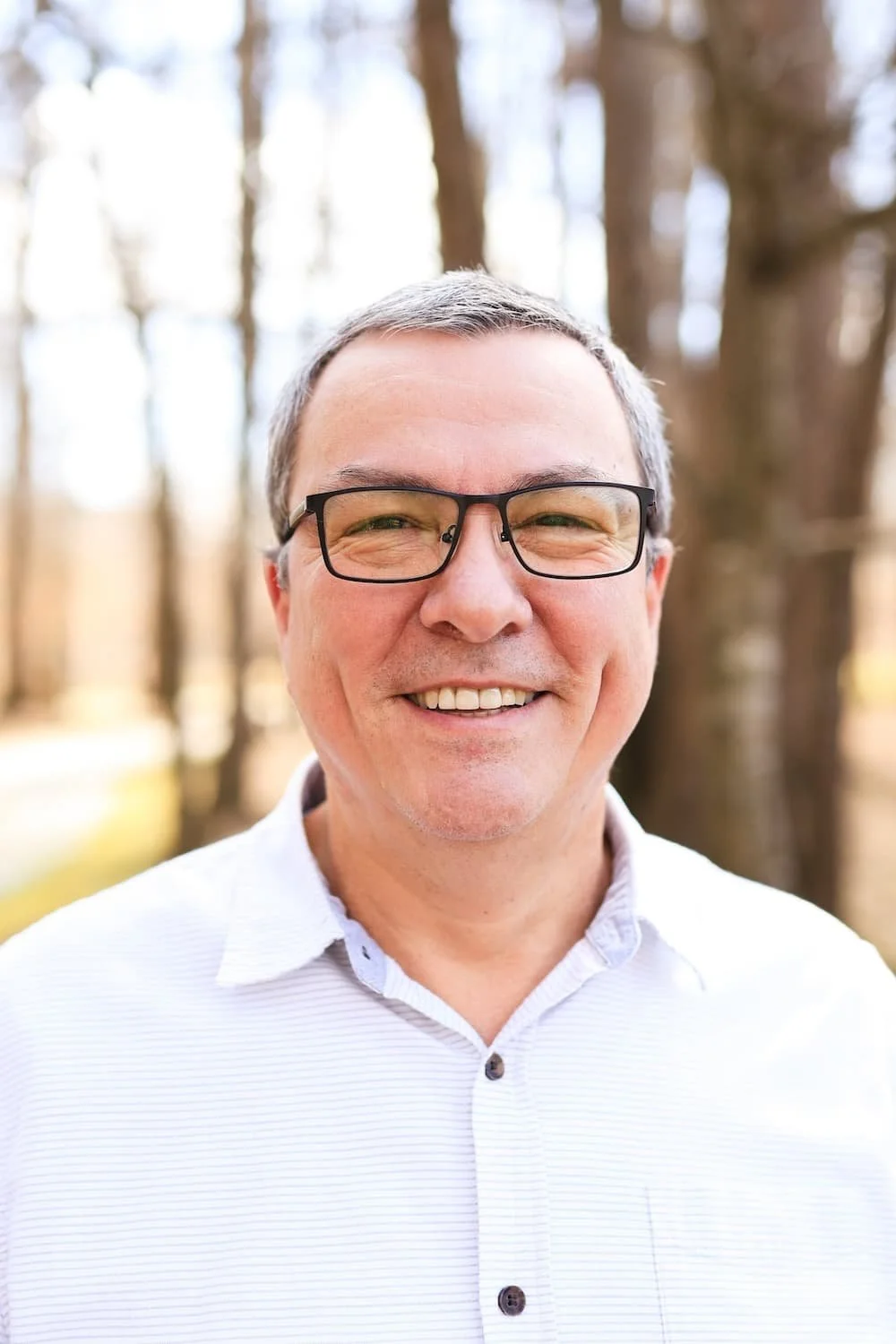 A middle-aged man with glasses and short gray hair smiling outdoors in front of a blurry wooded background in fall.