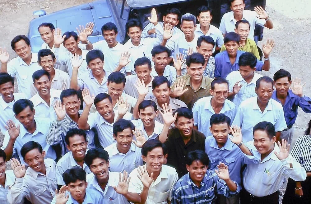 Group of smiling men waving at the camera, standing outdoors near a blue vehicle, with a dirt ground background.