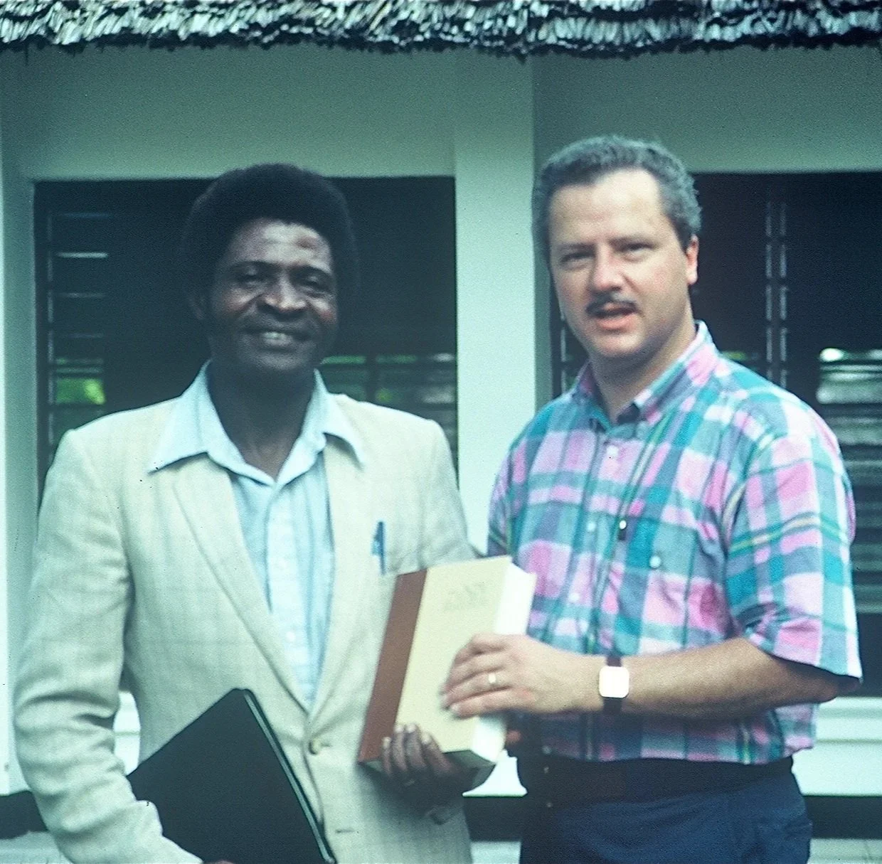Deux hommes debout ensemble devant un bâtiment, l'un tenant des papiers et l'autre un dossier, souriant à la caméra.