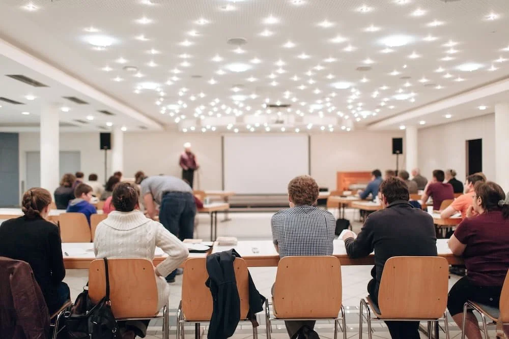 A conference room filled with attendees sitting at tables, facing a stage where a speaker is standing. The ceiling has numerous small bright lights.