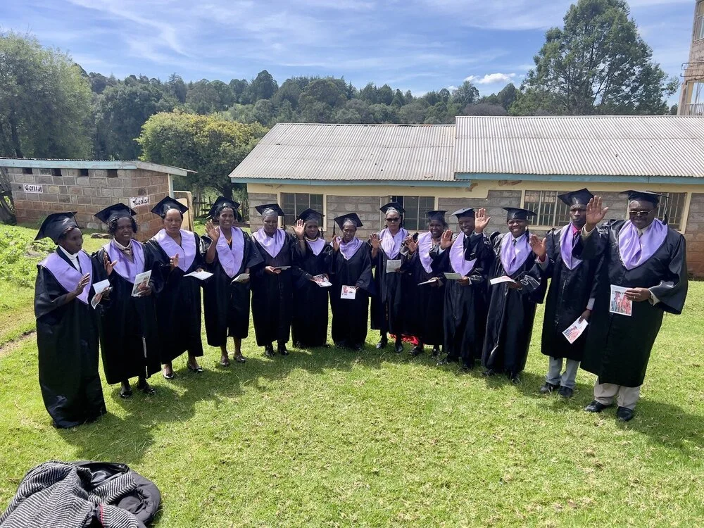 Group of students wearing graduation gowns and caps standing outdoors on a sunny day in Kenya, waving and holding books or pamphlets, with a building and trees in the background.