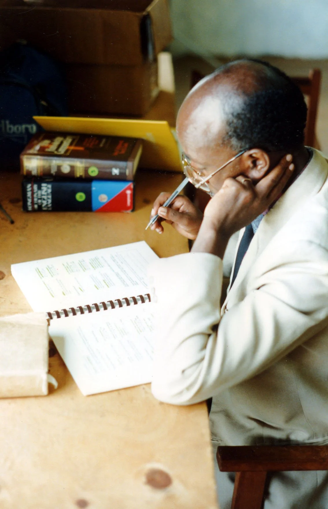 A man with glasses, wearing a light-colored suit, is seated at a wooden table, reading and writing in a book or notebook, with a pen in his hand. There are several books and a yellow folder on the table.