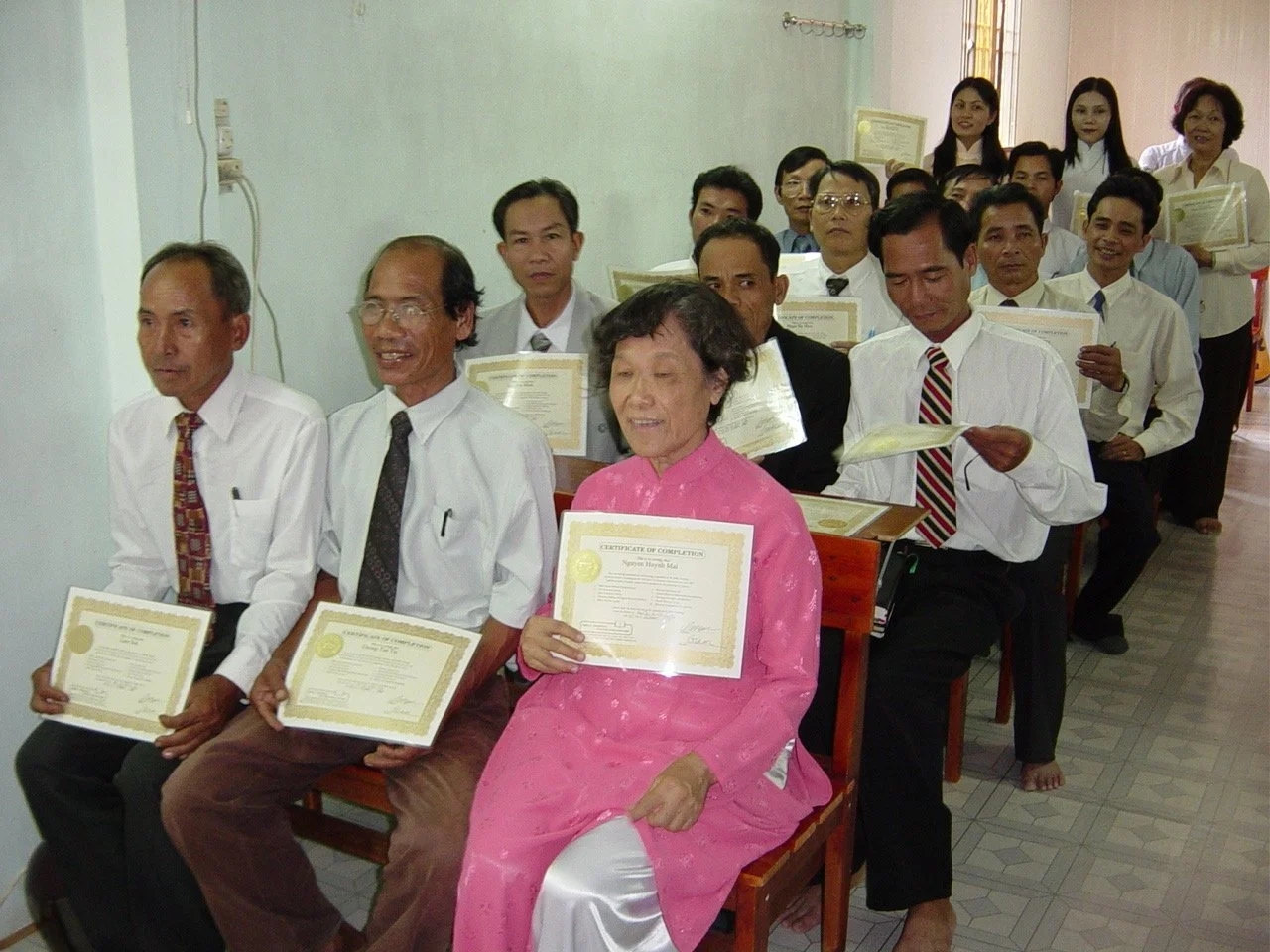 Group of adult men and women sitting in a room, holding certificates, at a formal event or graduation ceremony, with some standing in the background.