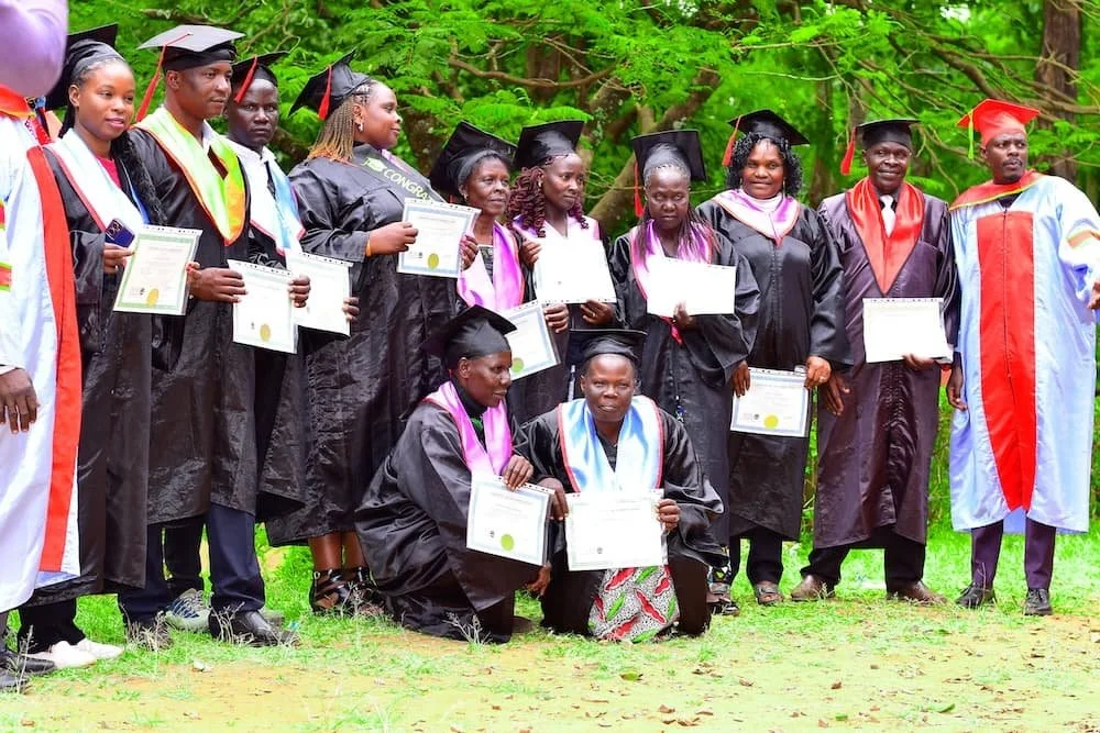 Group of graduation students wearing black caps and gowns, holding diplomas, standing outdoors on greenery background.