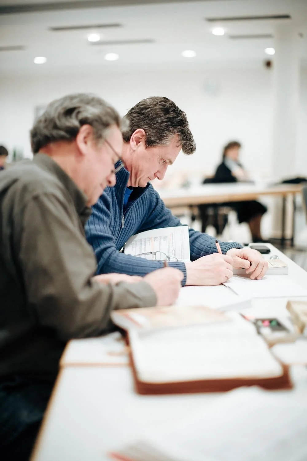Two men sitting at a table in a classroom, focused on writing notes during a Bible training session.