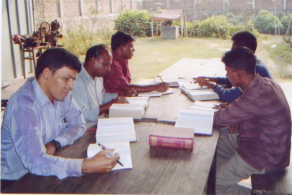 Five men sitting at a wooden table outdoors, reading and studying books, with a grassy garden and trees in the background.