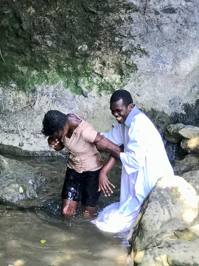 Dennis Mock, sitting at a table with a pen in his hand, looking directly at the camera, with a framed picture and a decorative metal heron on the wall behind him.