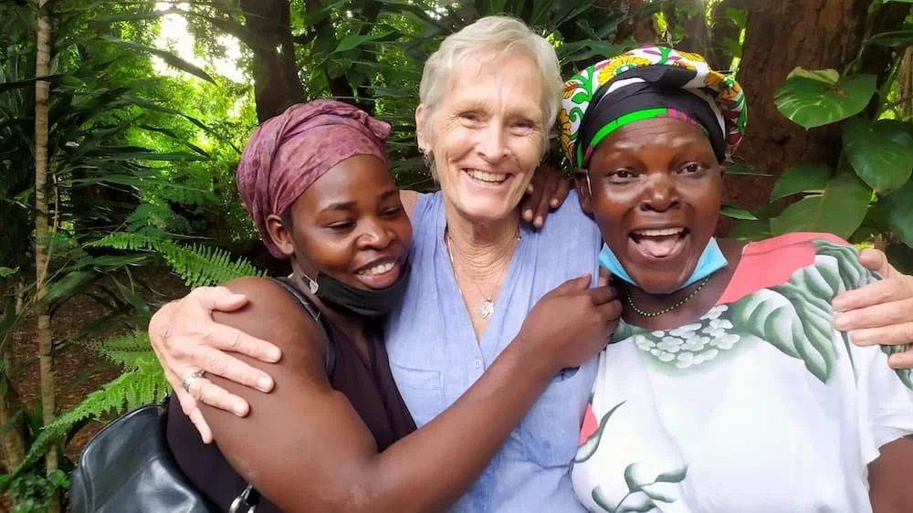 Trois femmes et une dame âgée, souriantes et enlacées, dans un cadre extérieur verdoyant, portant des foulards colorés et des vêtements décontractés.