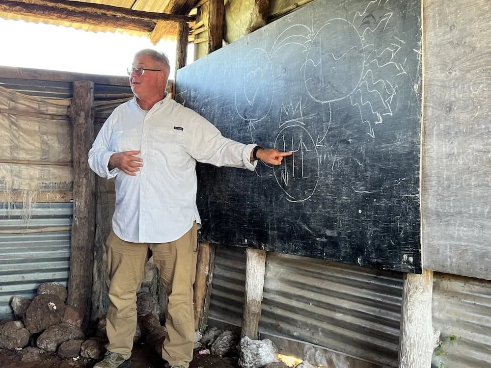 Un homme portant des lunettes et une chemise blanche boutonnée montre du doigt un tableau noir sur lequel sont dessinés des nuages et des montagnes. Il se tient debout dans un bâtiment rustique en bois et en métal, avec des pierres au sol.
