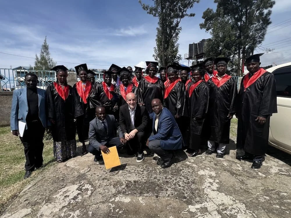 Group of graduates in black gowns with red stoles and mortarboards posing outdoors on a sunny day, with three men in suits kneeling in front.