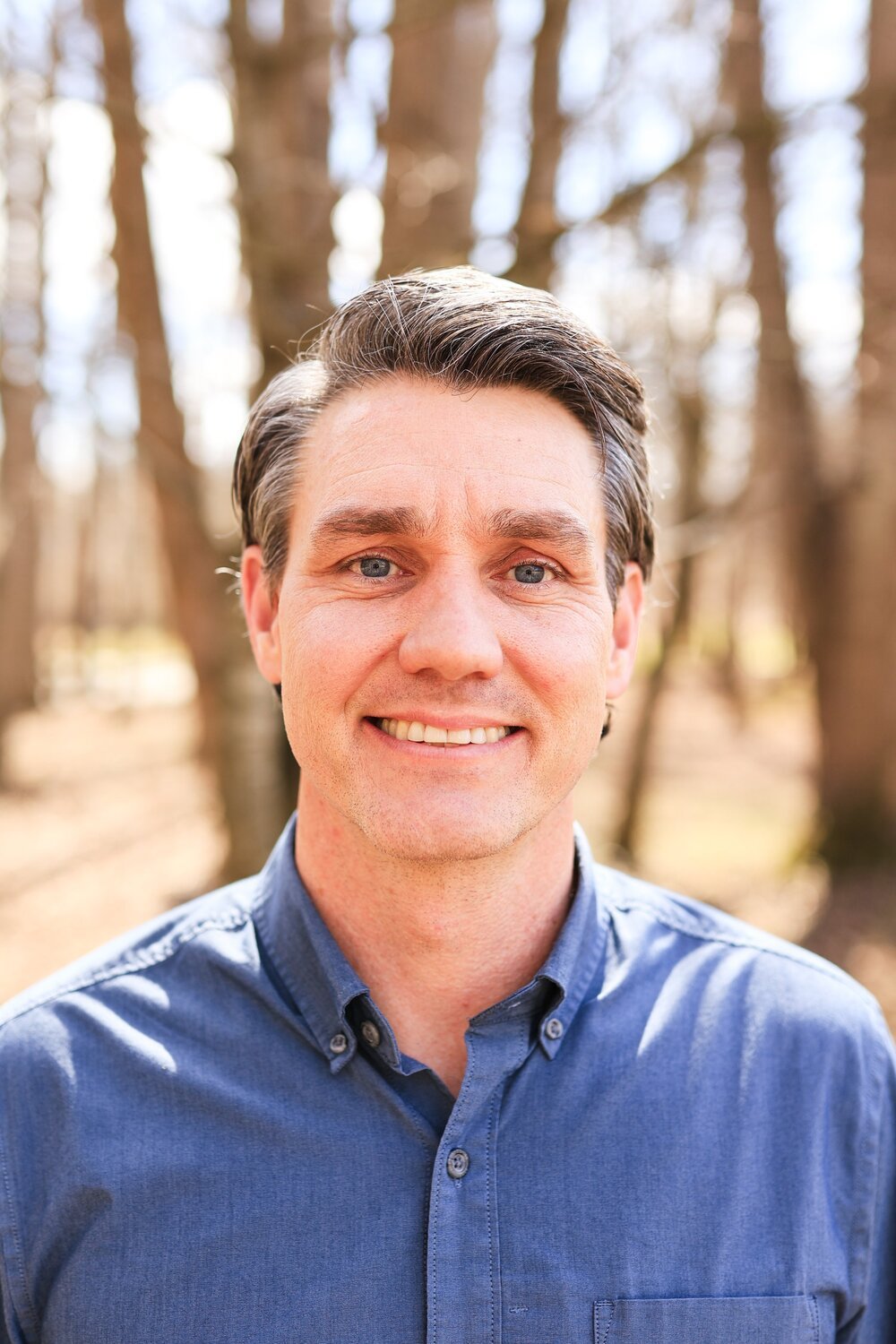 A smiling man with dark hair and blue eyes wearing a blue button-up shirt, standing outdoors in a wooded area with sunlight filtering through the trees.