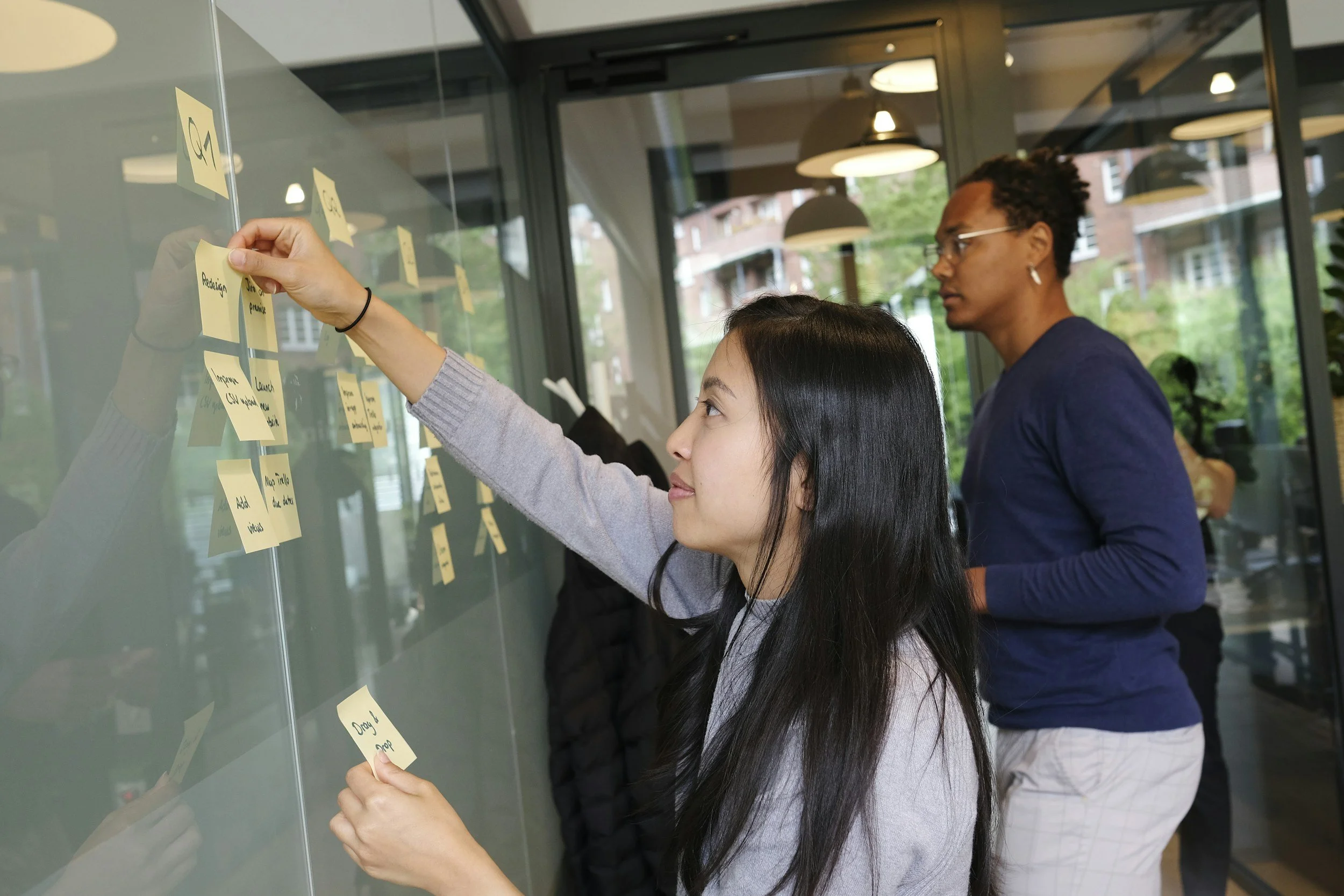 Two women are standing at a glass board with yellow sticky notes, one is placing a sticky note while the other looks on, in a modern office setting with large windows and ceiling lights.
