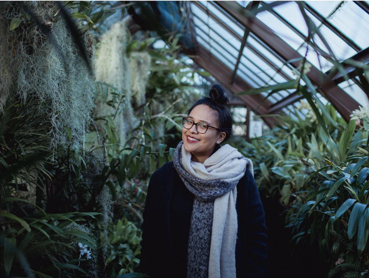 A woman with glasses smiling in a greenhouse surrounded by green plants and hanging moss.