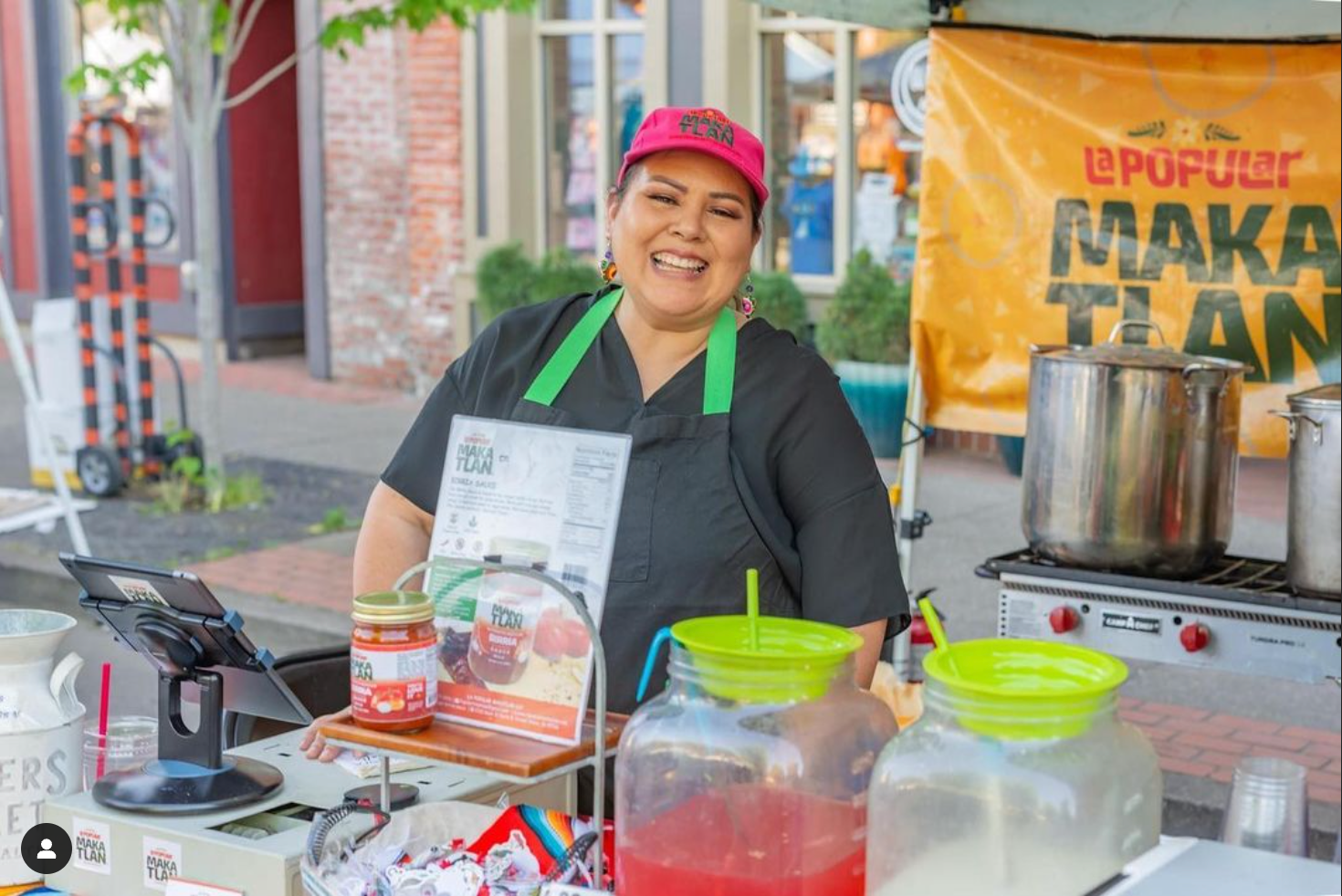 Smiling woman wearing a pink cap and black apron, standing at an outdoor food stand with jars, containers, and a large pot, promoting Makatán food.