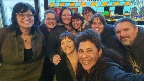 A group of eight smiling people standing closely together inside a colorful indoor setting.