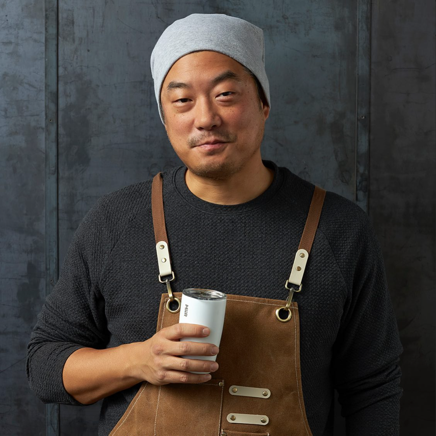 A man with a gray beanie and black sweater wearing a brown apron holding a white tumbler with a lid, standing in front of a dark textured metal wall.