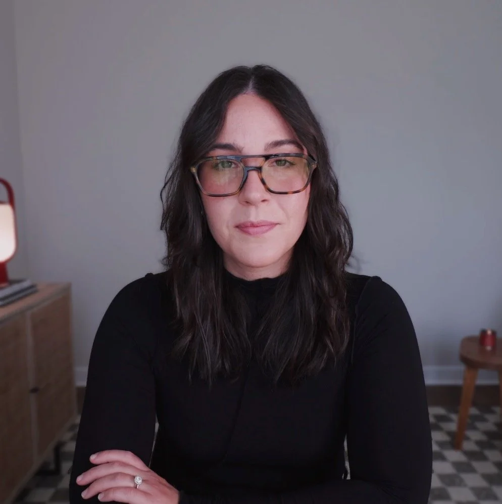 A woman with wavy dark hair and glasses, wearing a black top, sitting in a room with a gray wall, wooden side table, and checkered floor.