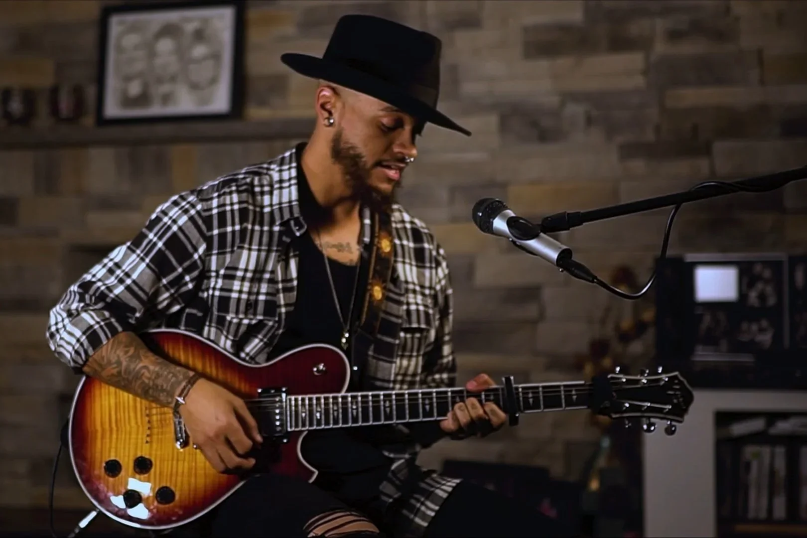 A man in a black hat, plaid shirt, and tattoos playing an electric guitar in front of a microphone in a room with wood paneling.