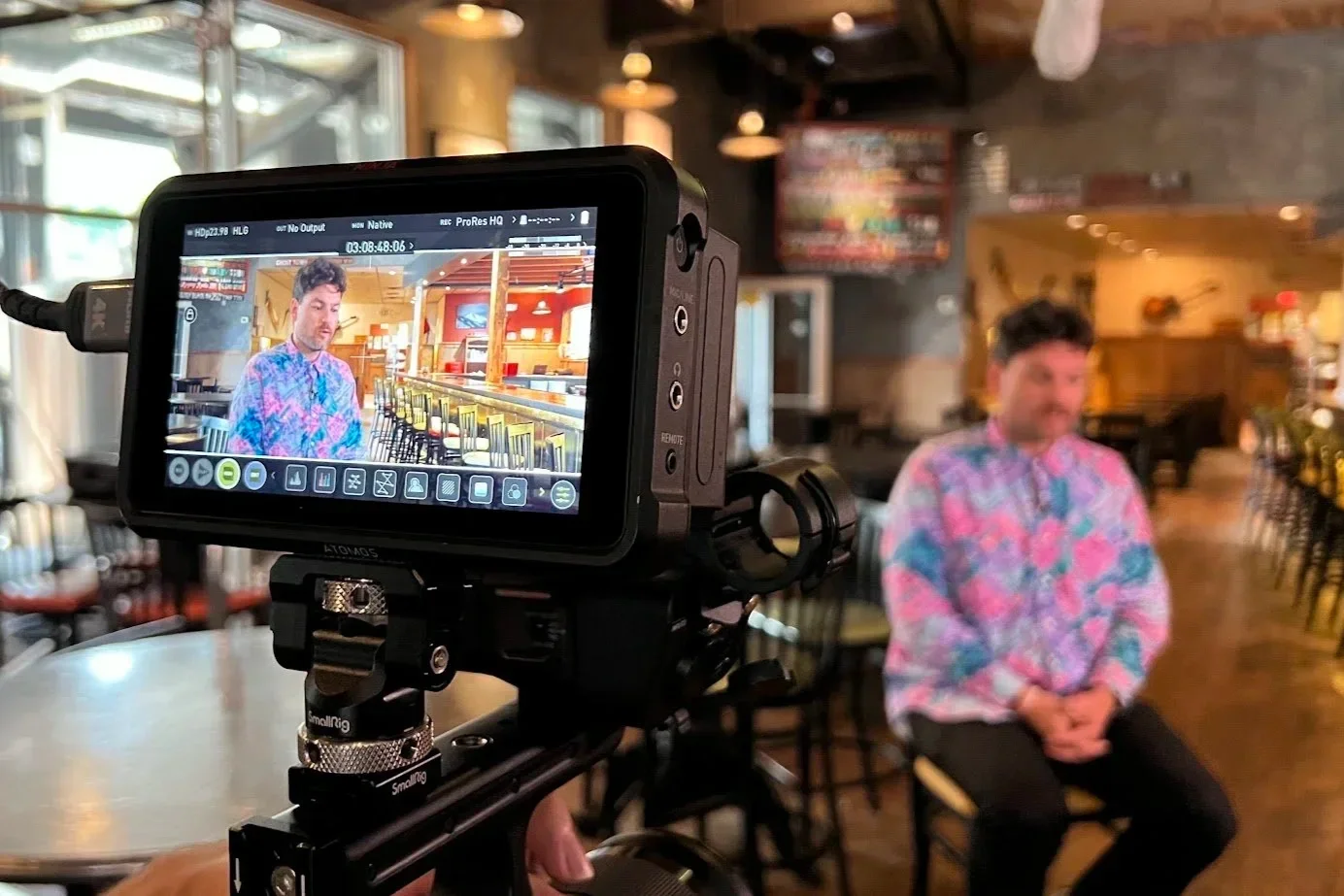 A man in a colorful, patterned shirt is sitting on a chair in a dimly lit restaurant or bar, while a professional camera on a tripod records him. The background shows tables, chairs, and walls decorated with musical instruments and artwork.