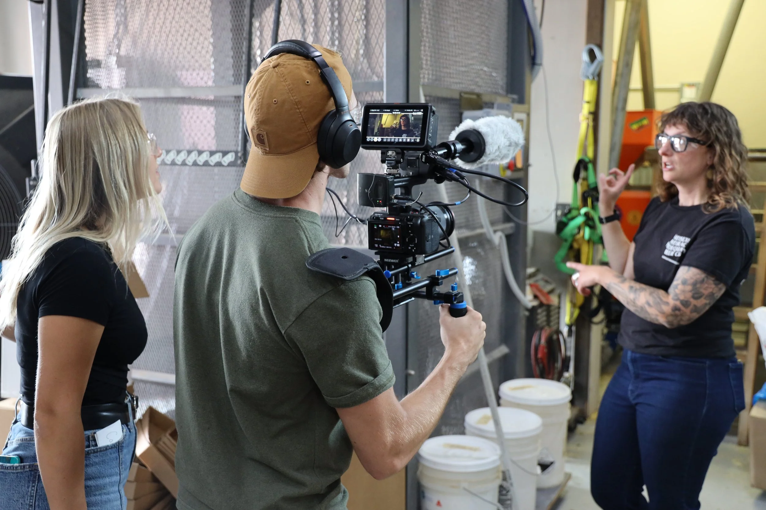 A woman with tattoos on her arm is speaking to a camera crew in a workshop. The woman is gesturing with her right hand as she talks, and the crew is recording her with a professional camera and a person wearing headphones. Two other women are standing nearby, one with blonde hair in a black shirt and the other with long blonde hair, glasses, and a black shirt.