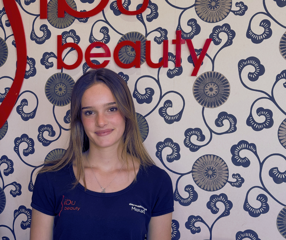 A young woman with shoulder-length brown hair smiling softly in front of a decorative background with navy blue floral patterns and large red text that reads 'ipU beauty'.