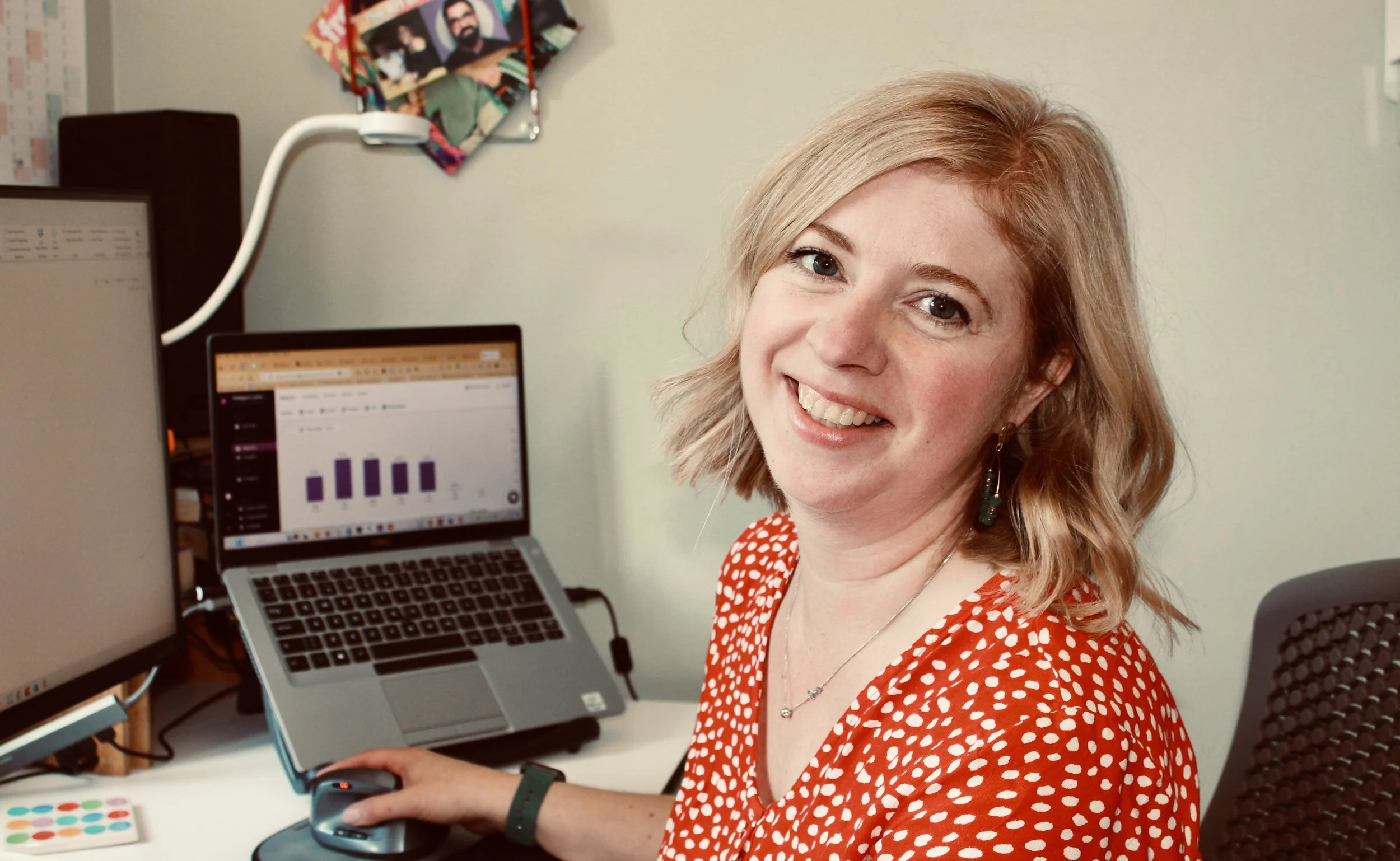 A woman with blonde hair and earrings sitting at a desk working on a laptop, with office supplies and a computer monitor displaying a bar graph.