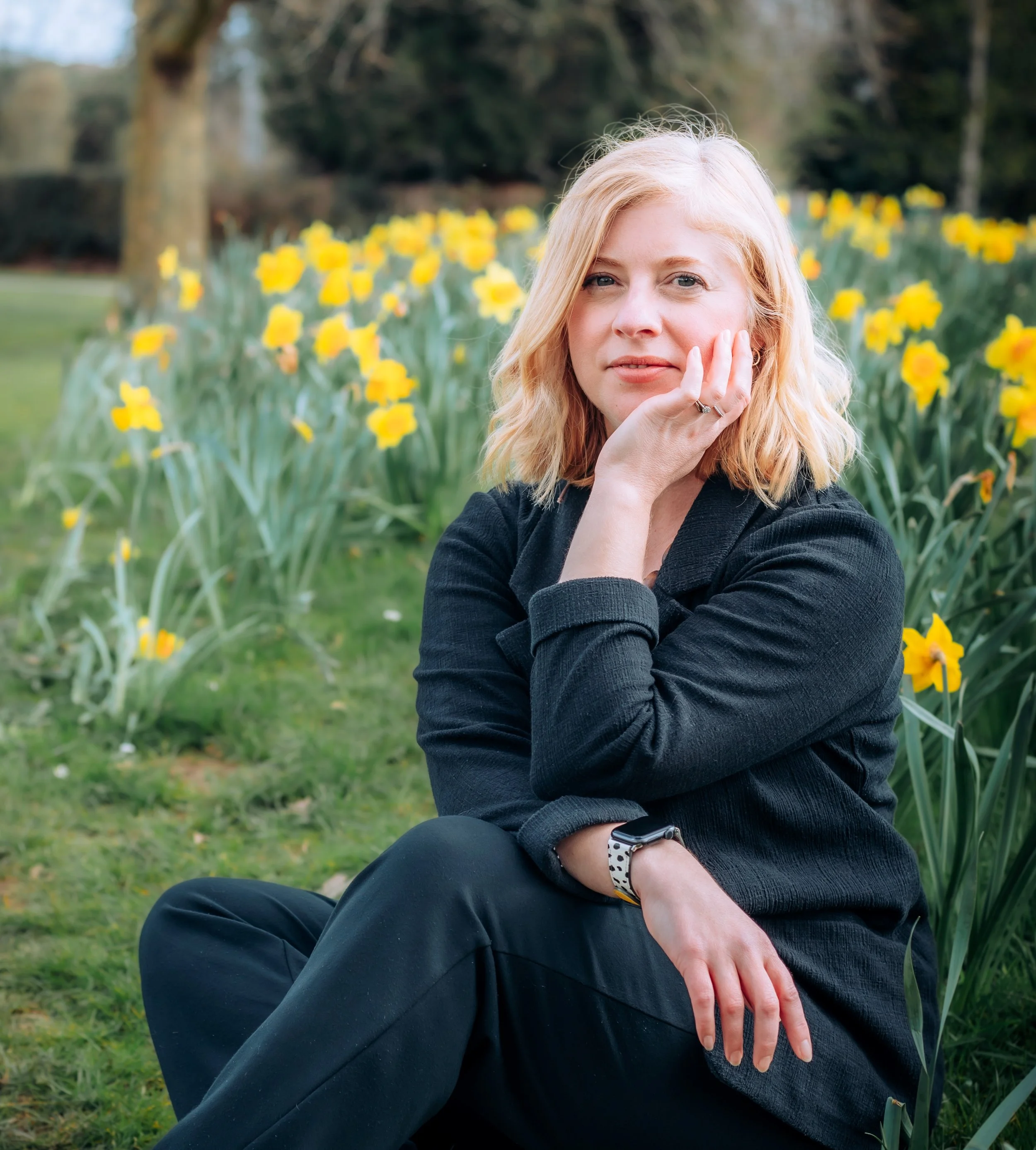 A woman with blonde hair wearing a black jacket sitting on grass with yellow flowers in the background.