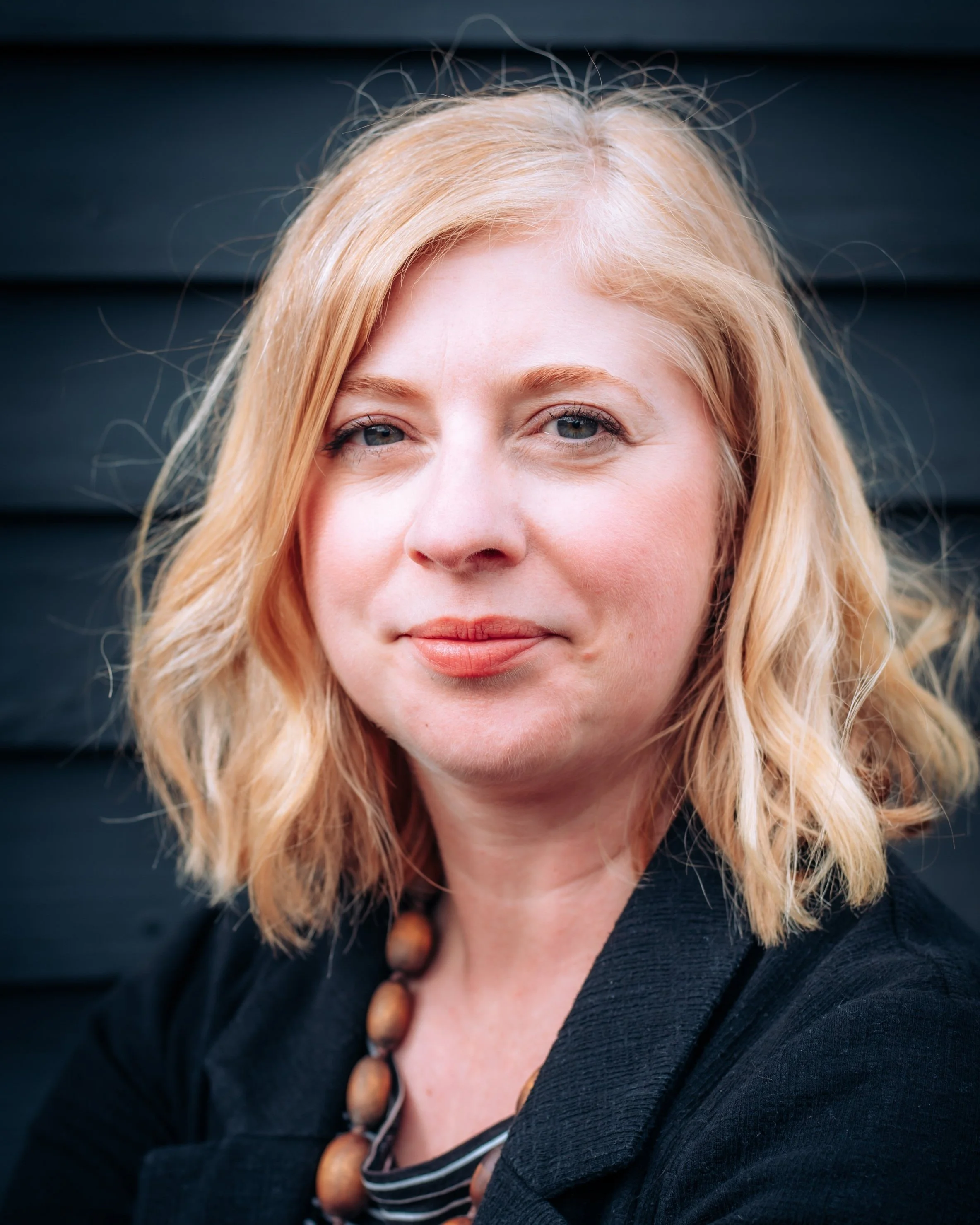 Portrait of a woman with blonde hair, blue eyes, and red lipstick, wearing a black jacket and a wooden bead necklace, standing in front of a dark wooden wall.