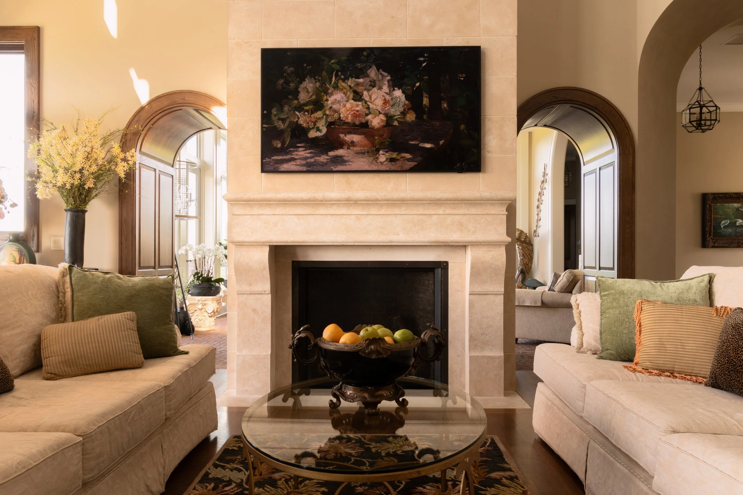 Elegant living room with a large stone fireplace, floral painting above, beige sofas with decorative pillows, a glass coffee table with fruit bowl, and archways leading to other rooms. Bright and spacious interior design.