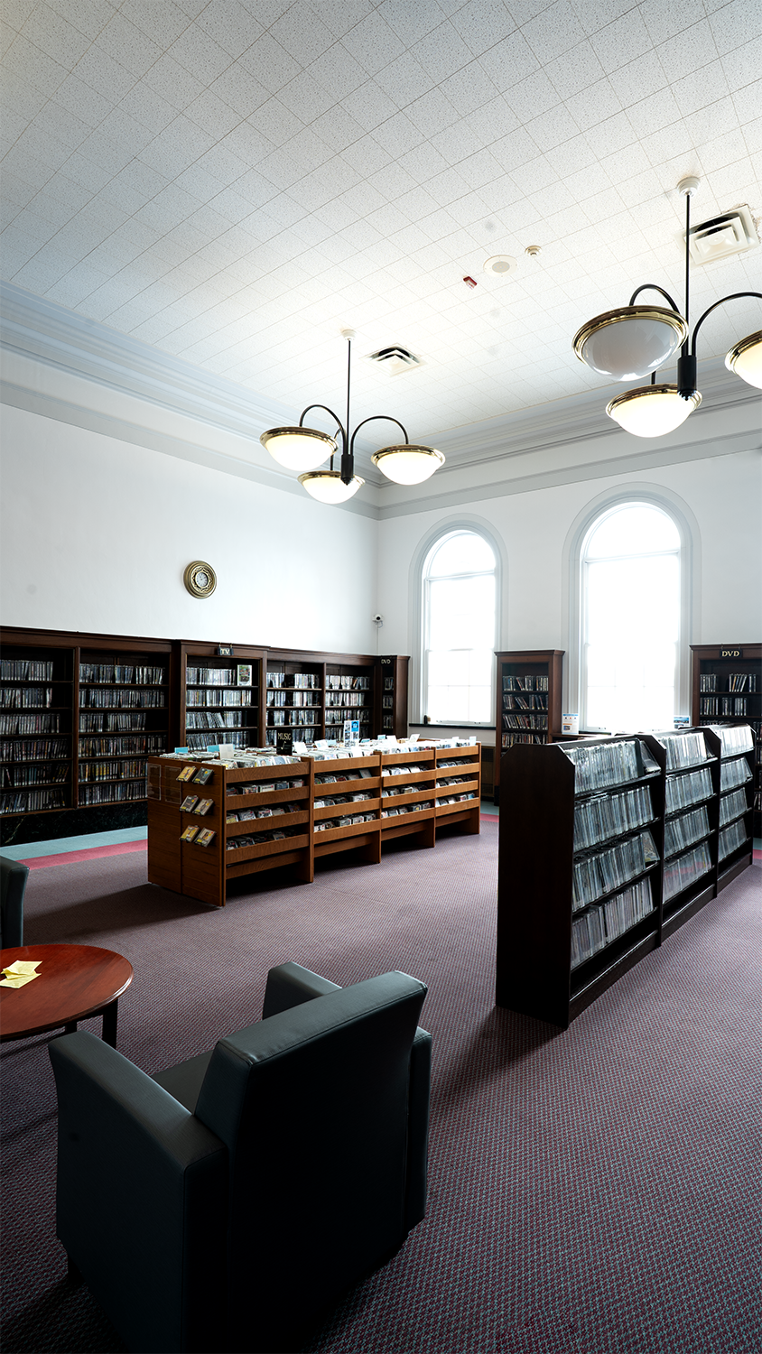 Library interior with bookshelves, CDs, DVDs, armchairs, large windows, and ceiling lights.