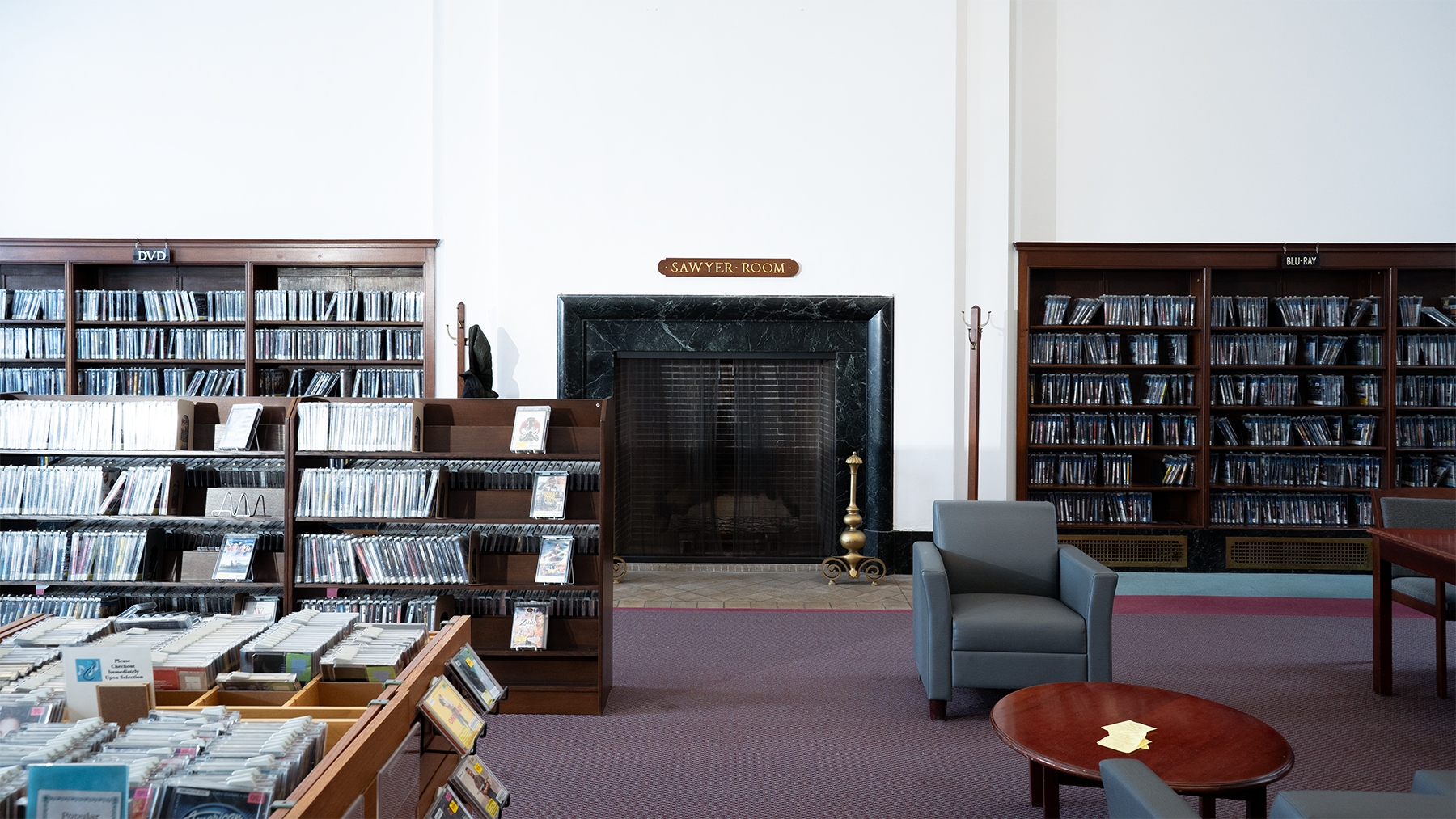 Library media room with shelves of DVDs and Blu-rays, a fireplace labeled 'Sawyer Room,' seating area with chairs and a table.