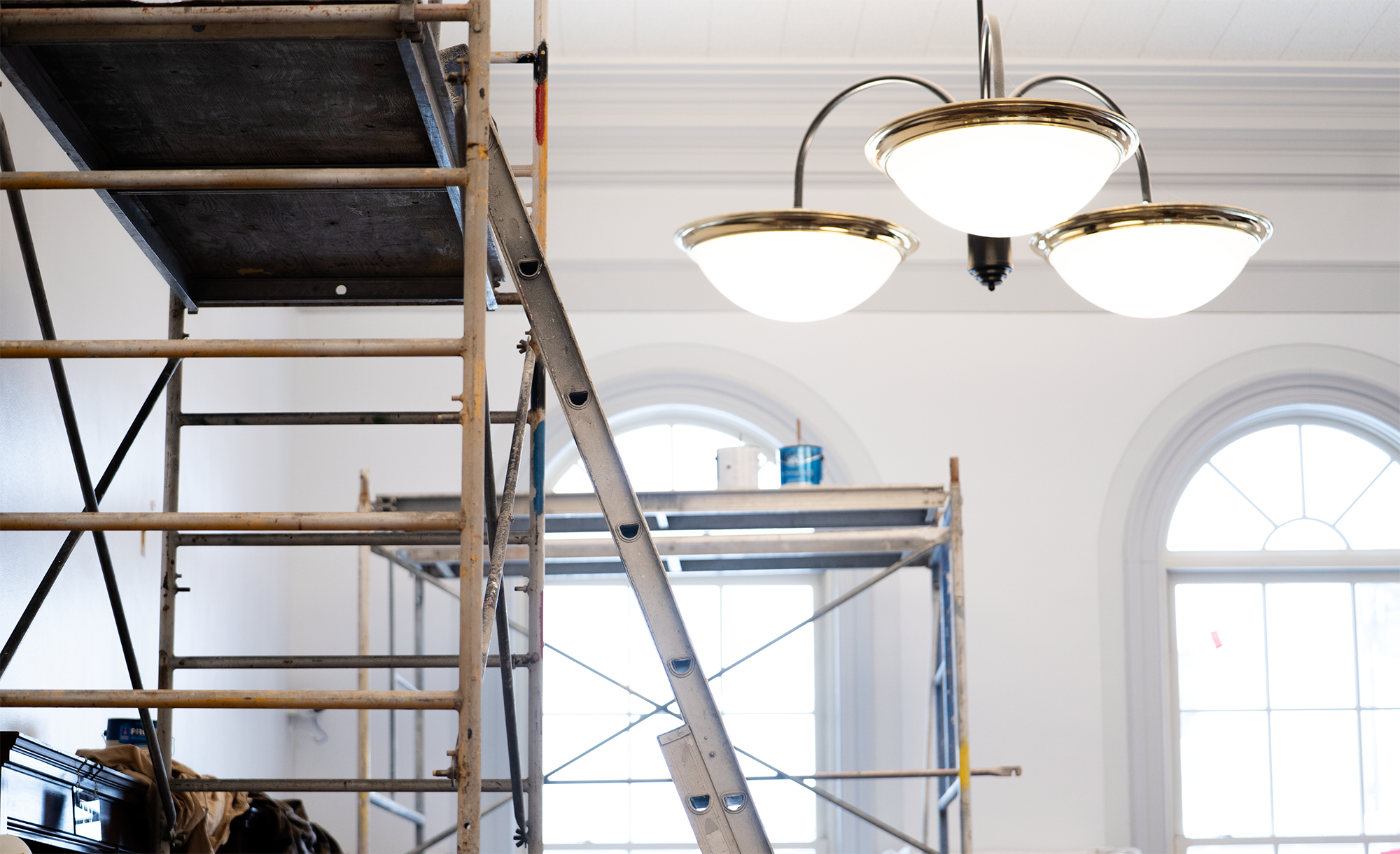 Indoor renovation scene with scaffolding, ladder, and hanging light fixture, featuring arched windows in the background.
