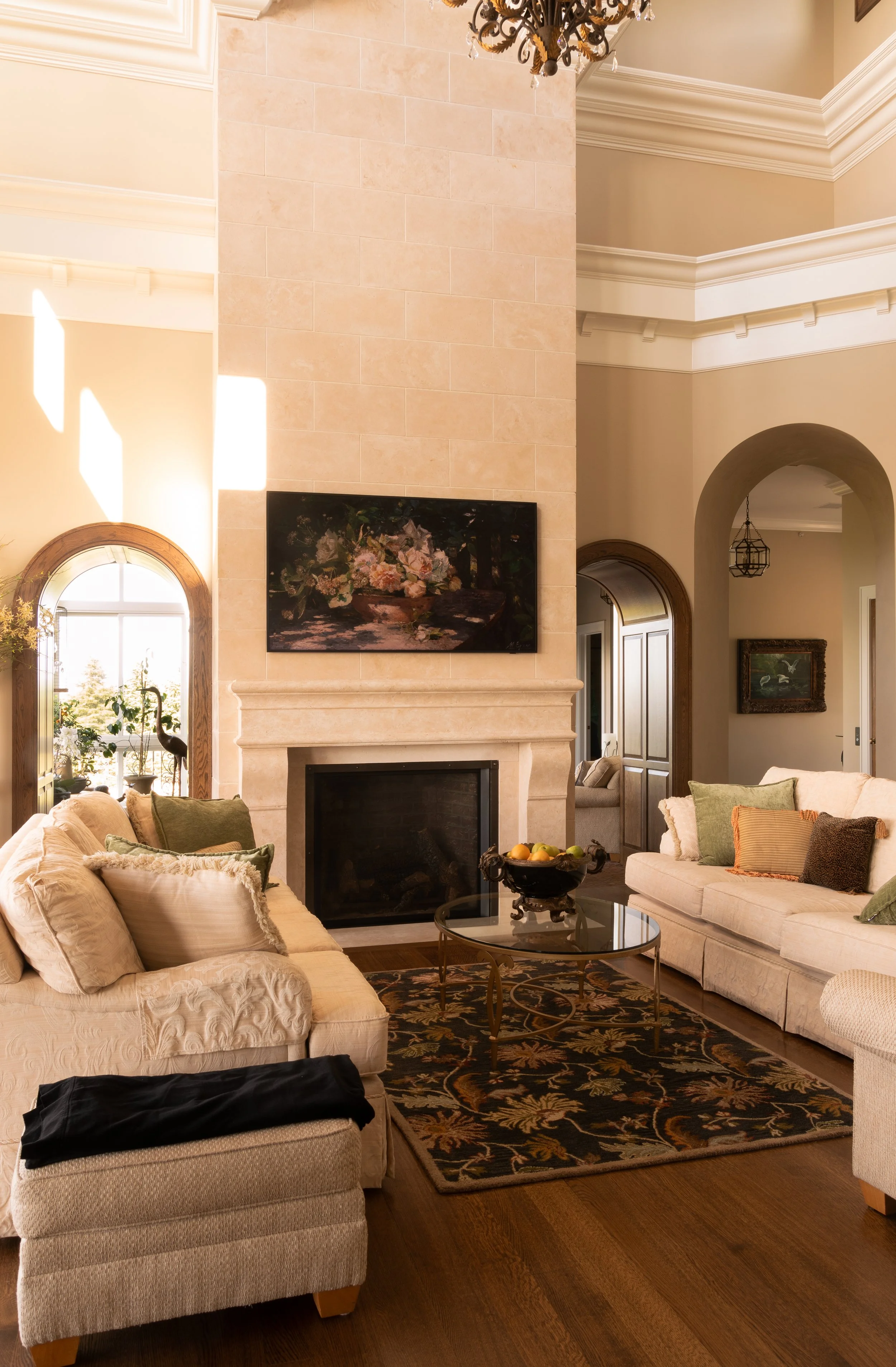 Elegant living room with high ceiling, fireplace, floral painting, beige couches, decorative pillows, glass coffee table, fruit bowl, patterned rug, and large windows.
