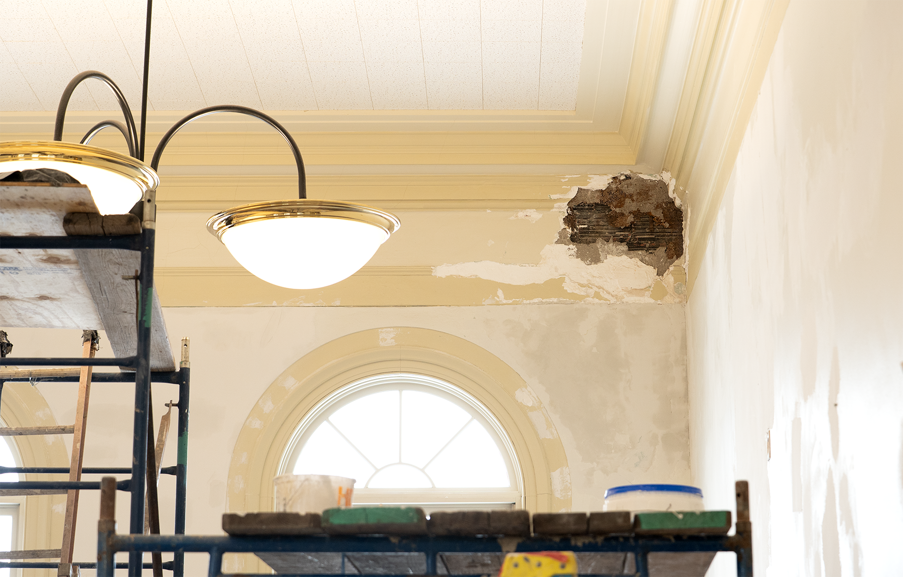 Interior room under renovation with scaffolding, ceiling light fixture, and a damaged wall with plaster and exposed lath near a window.