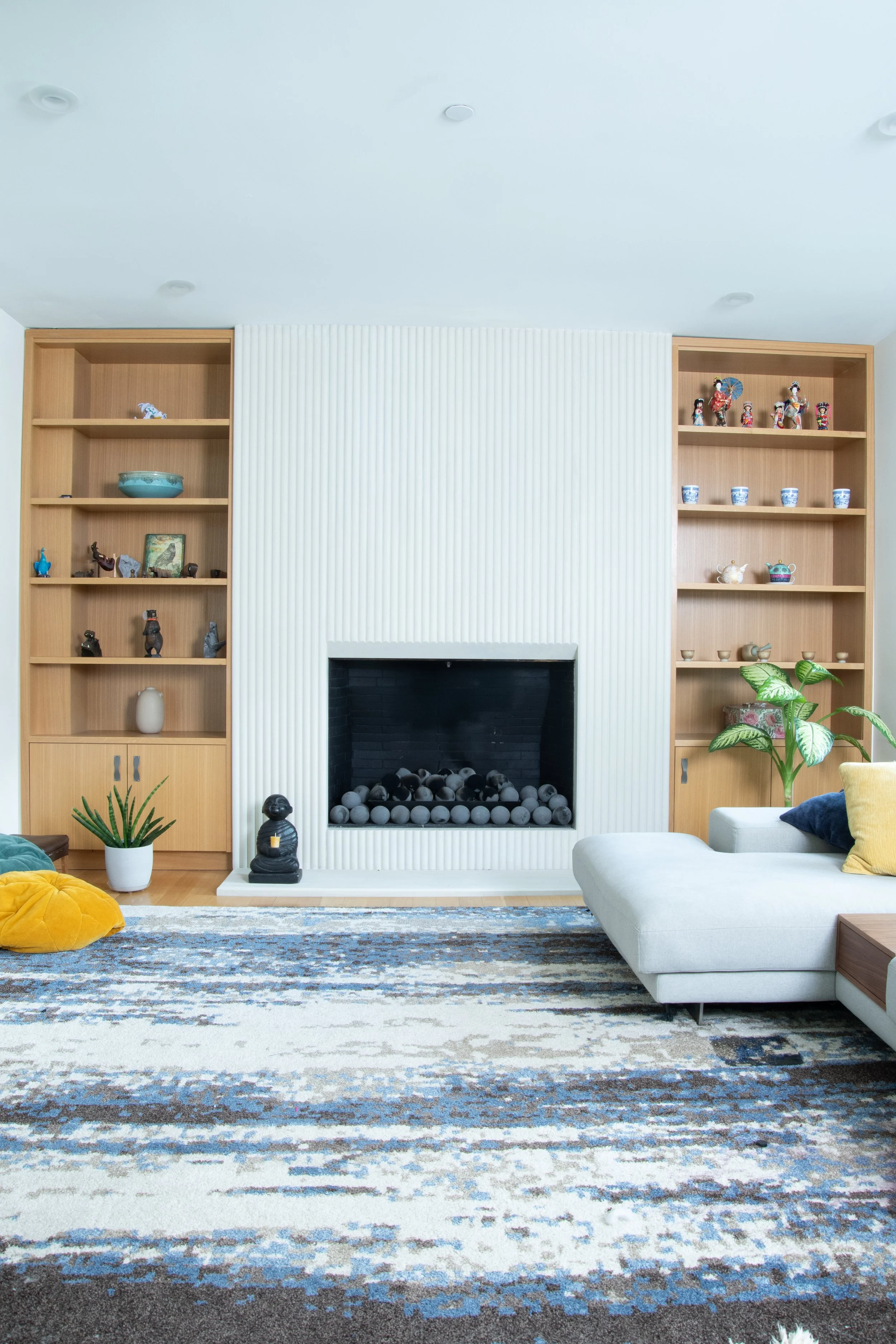 Modern living room with a minimalist design featuring a central fireplace, flanked by wooden shelves displaying decorative items. A large, abstract-patterned rug covers the floor, and a light gray sectional sofa with colorful cushions is positioned to the right. A small plant sits on the left near a round, mustard-yellow pillow. The space is bright and airy, emphasizing open, clean lines.