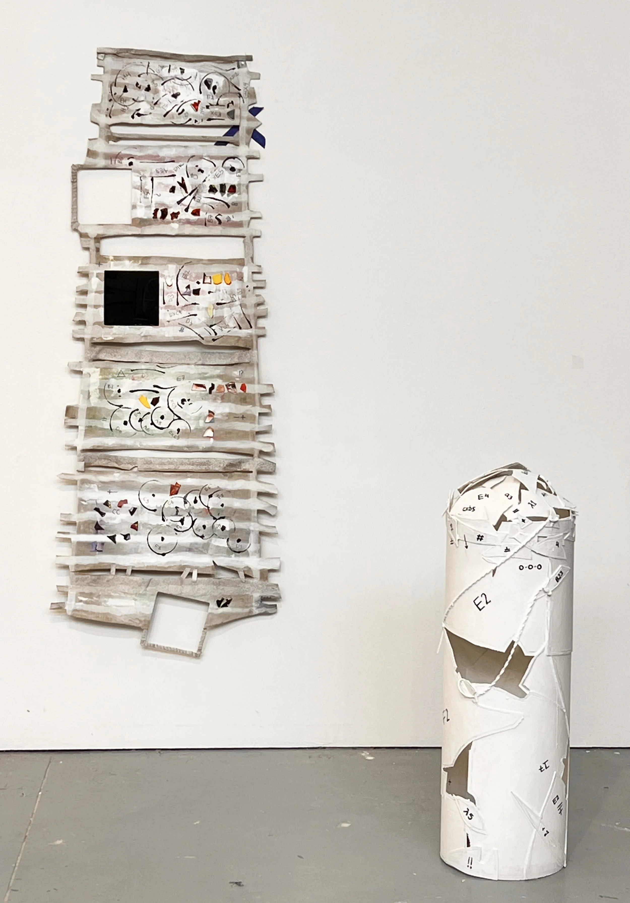 Mixed media art installation featuring a tall, narrow framed piece on the left with various abstract shapes and sections, and a white cylindrical sculpture on the right with torn paper and printed markings.
