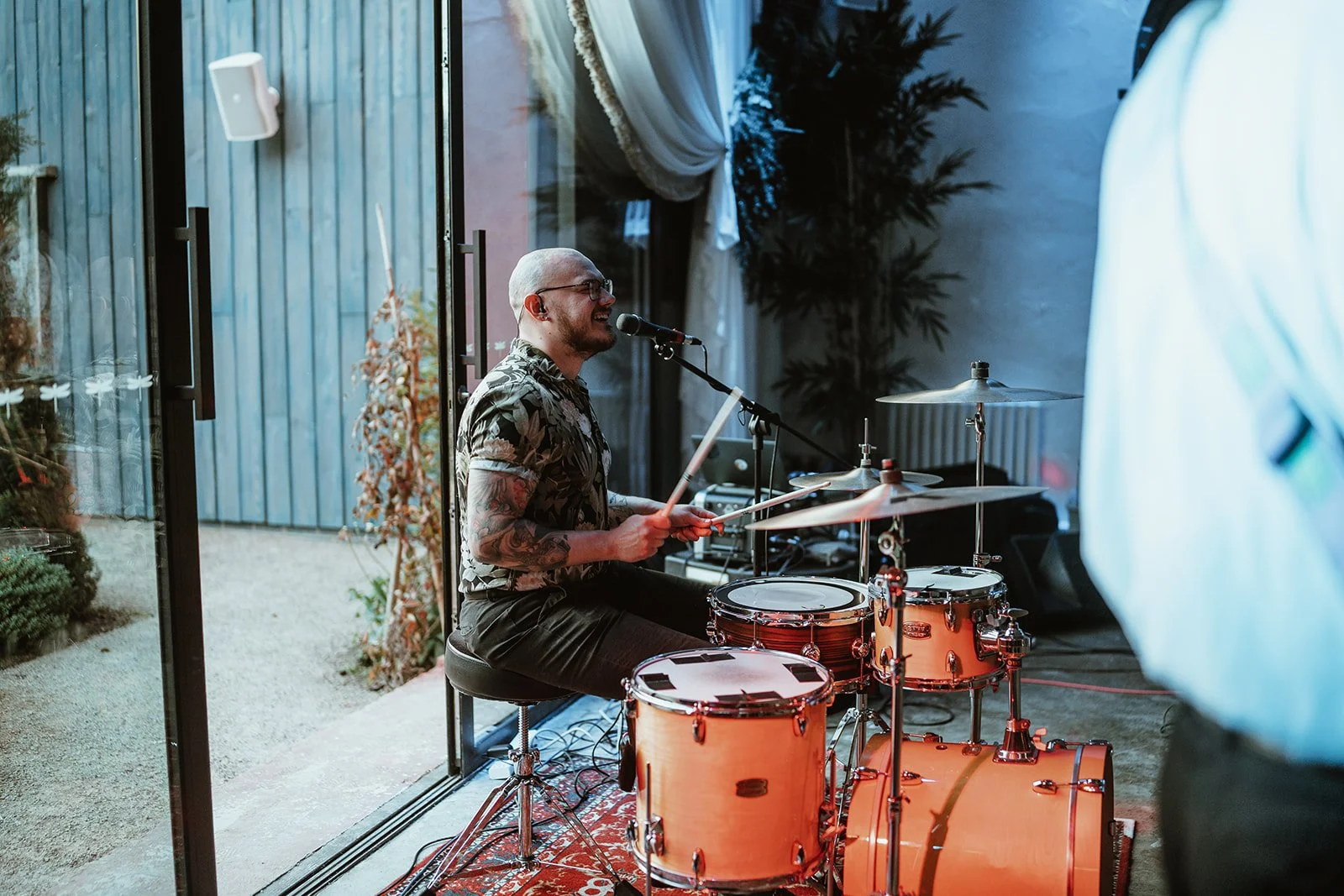 A man with tattoos and glasses singing and playing drums indoors near a glass door leading outside - Runa Farm North East wedding band - book the best wedding band - the Gatenby's - Newton Hall 
