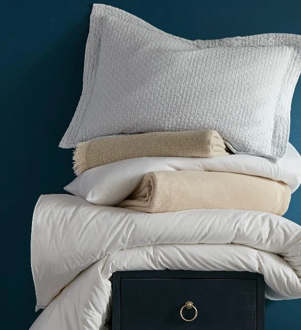 Stack of neatly folded bedsheets and blankets topped with a textured gray pillow on a bedside table against a dark teal wall.