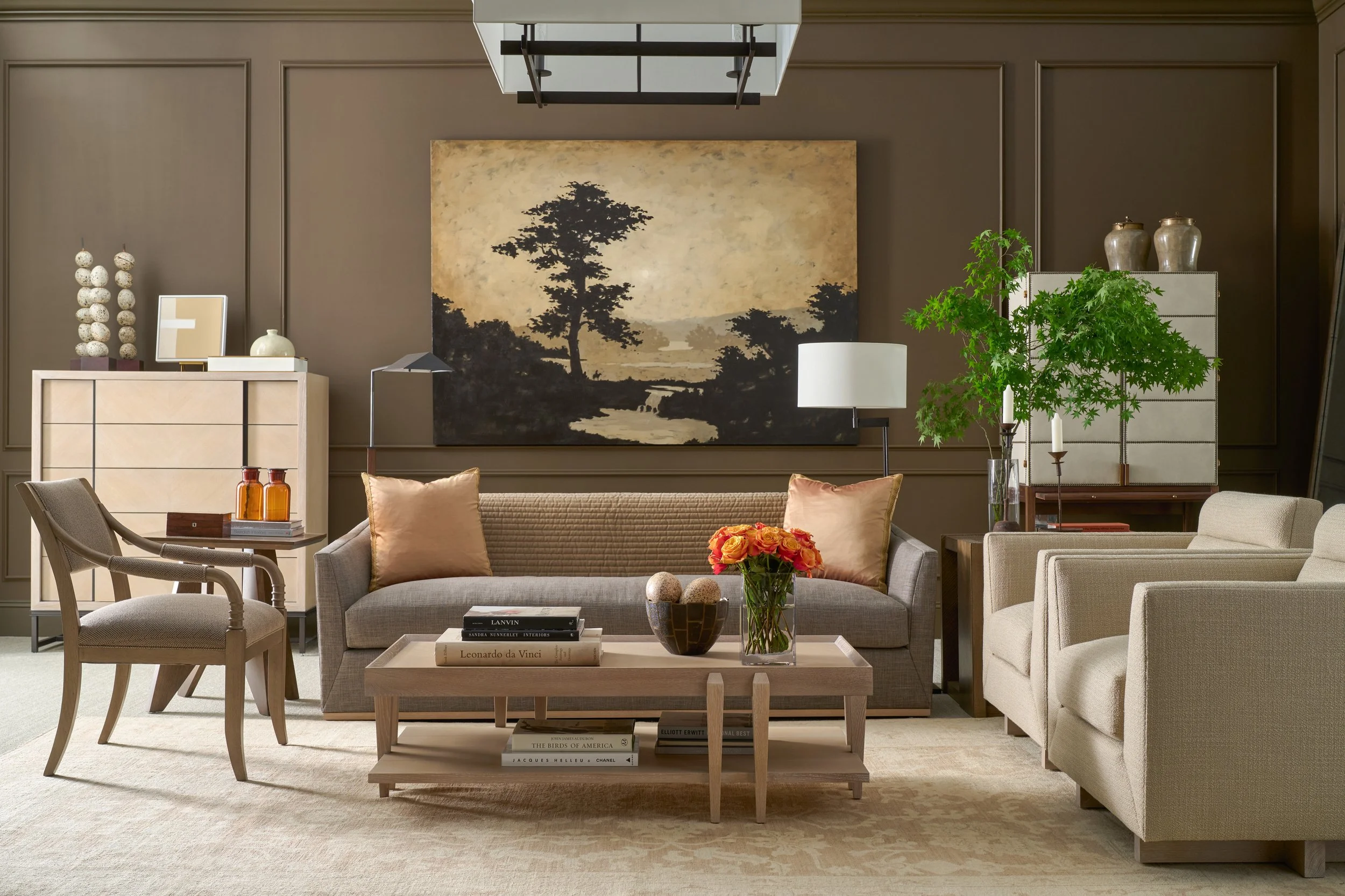 Elegant living room with beige and taupe furniture, a large landscape painting of trees on the wall, a coffee table with books and a flower vase, and a green plant in a glass vase.