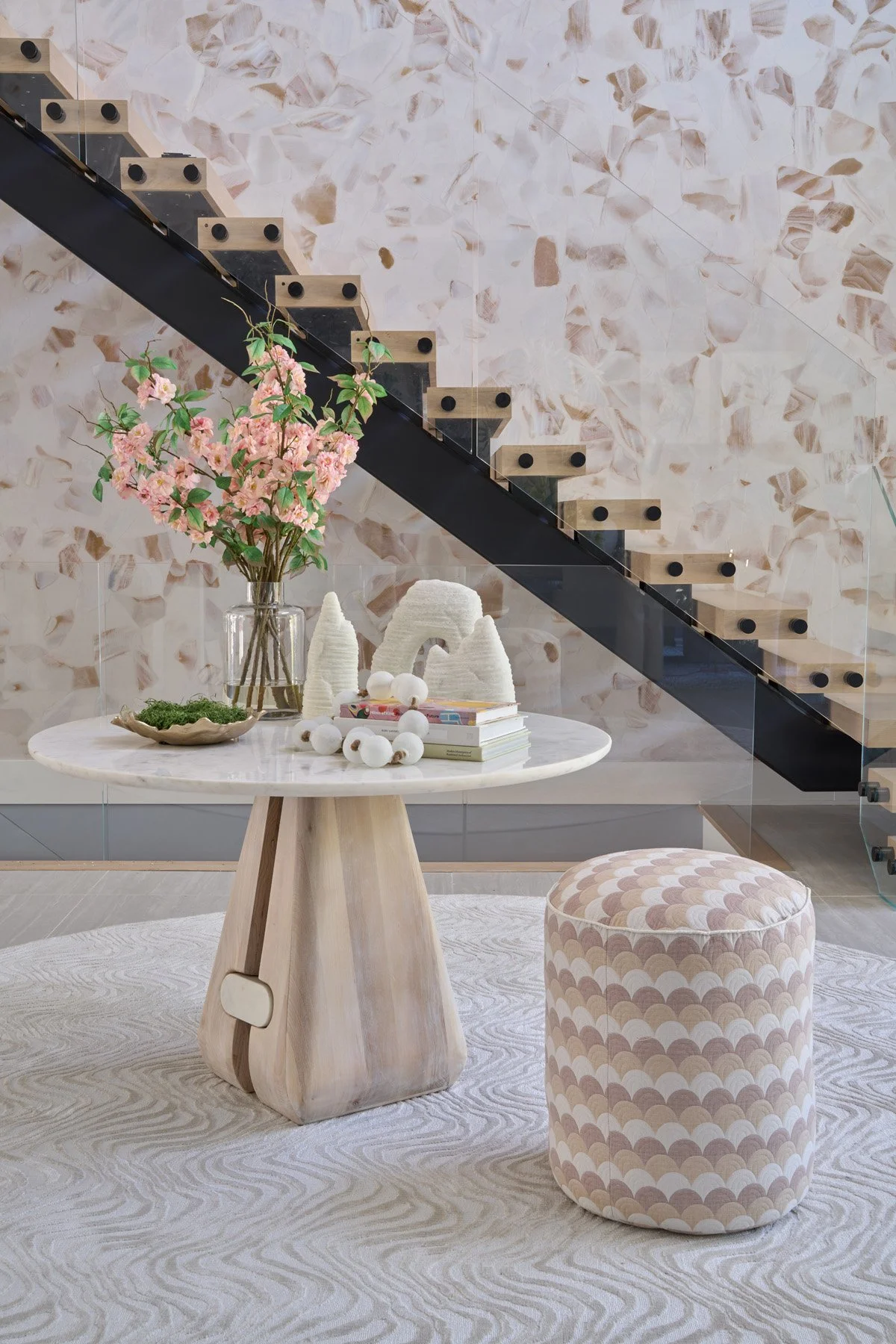 Interior of a modern living space featuring a round table with floral arrangements and decorative items, a pink and beige patterned ottoman, and a staircase with wooden steps and a black railing against a textured pink wall.