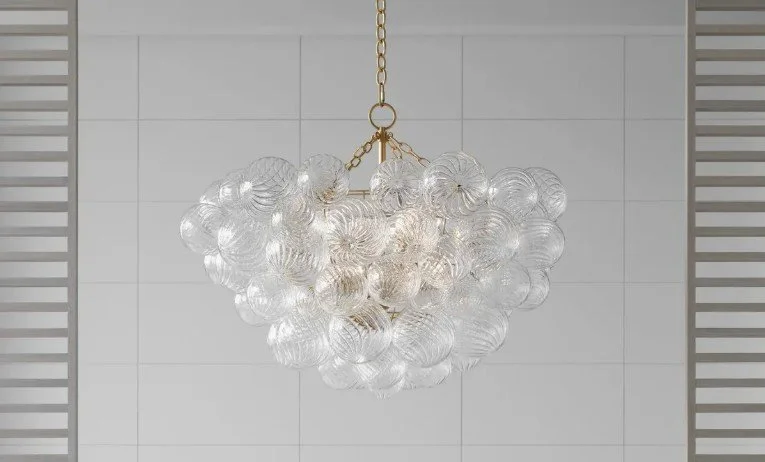 A modern chandelier composed of multiple clear glass globes hanging from a gold chain, set against a white tiled wall background.