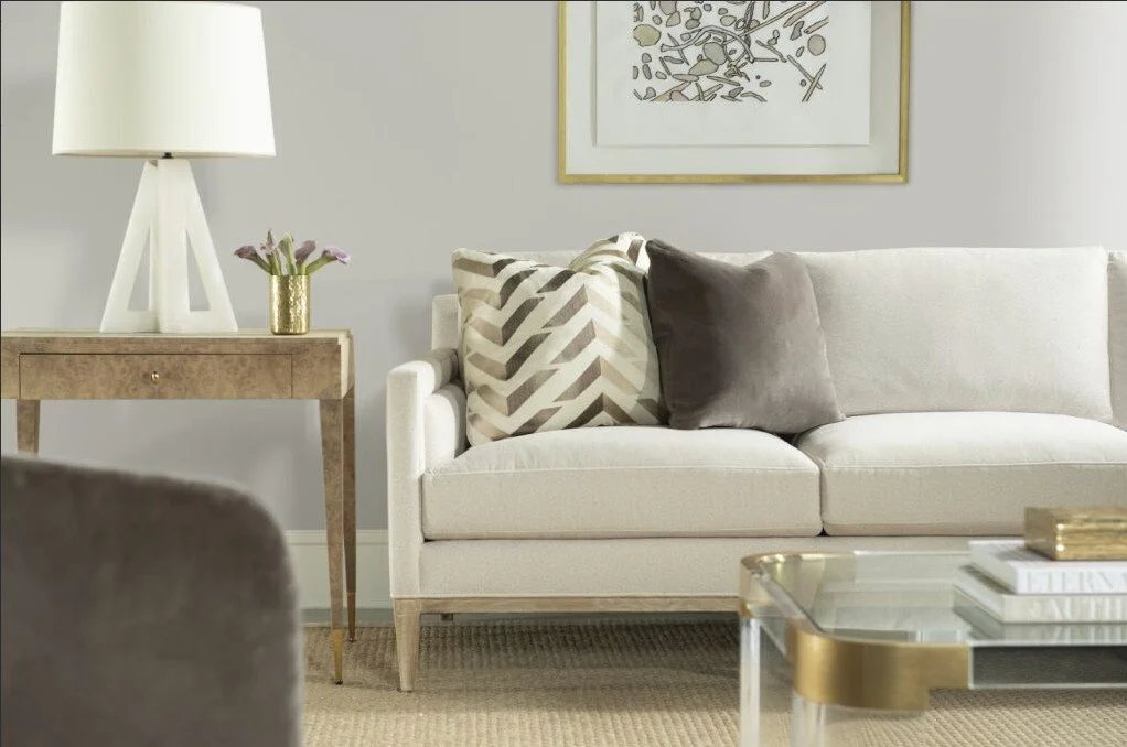 A beige living room with a white sofa decorated with two pillows, one patterned and one plain. A wooden side table with a white lamp and a small vase of purple flowers, and a glass coffee table with books. Wall art is mounted above the sofa.