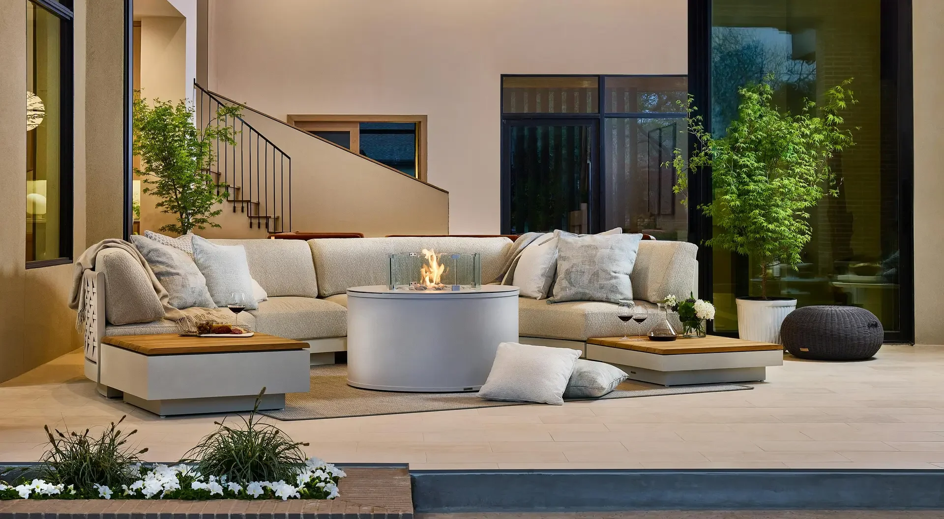 Modern outdoor living space with a sectional sofa, a round fire pit, plants, and large glass doors.