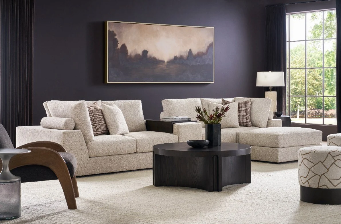 Living room with beige sectional sofa, black coffee table, artwork, and large window showing greenery outside.