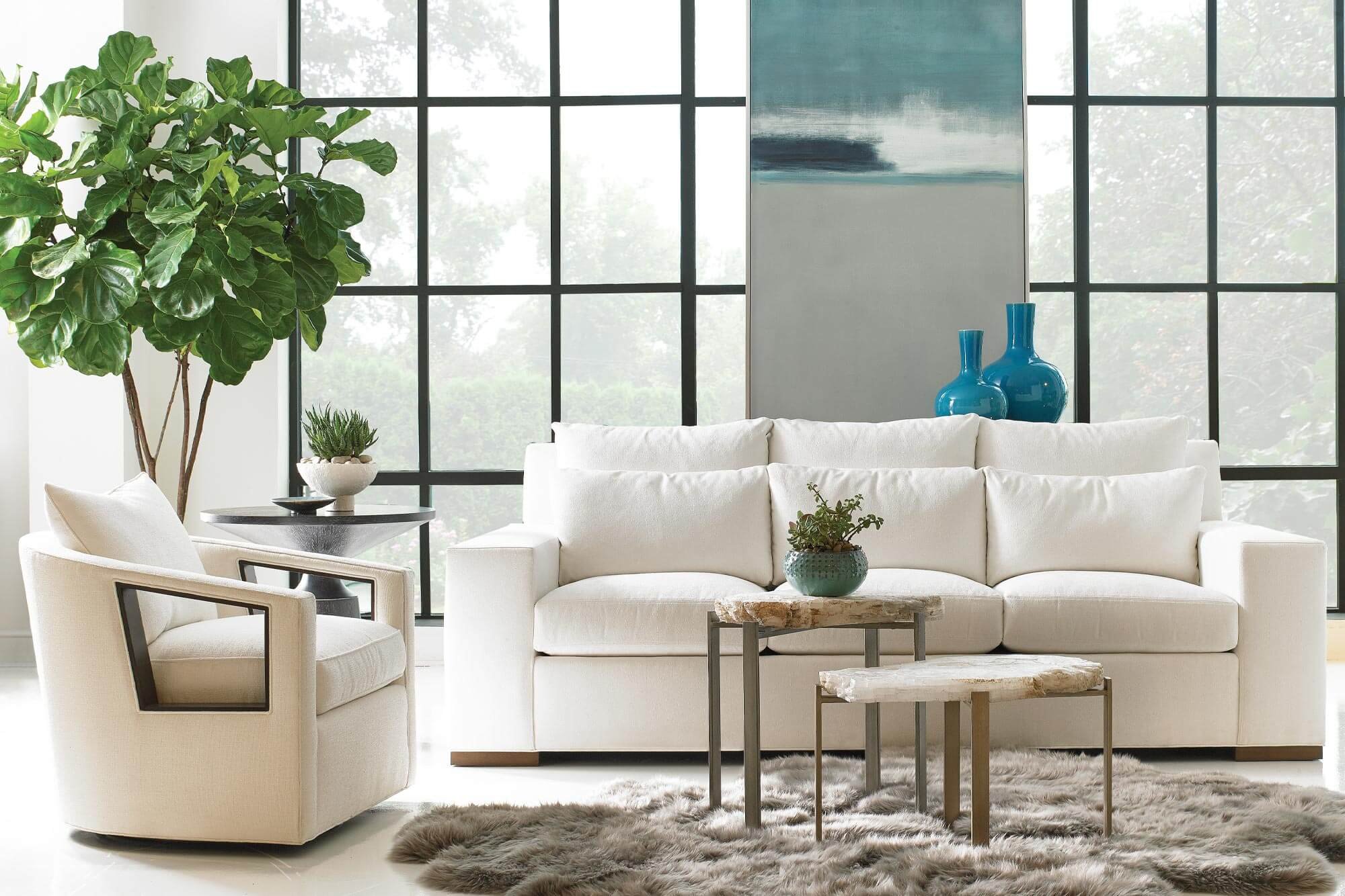 Living room with a large white sofa, a matching white armchair, a gray shag rug, and a large green plant in the corner. There are two small wooden tables with metal legs in front of the sofa, and decorative vases and potted plants on the tables and against the window. The room has large grid windows with a view of trees outside and an abstract painting on the wall.