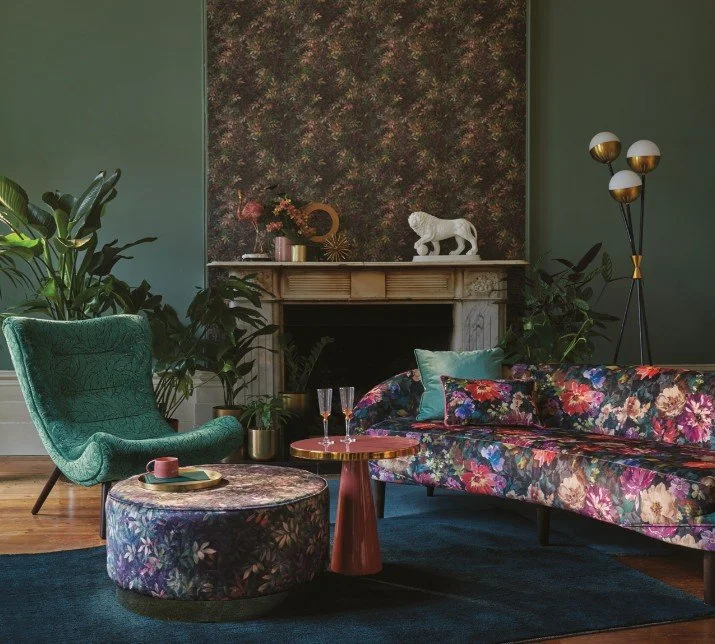 A living room with green walls, a floral wallpaper accent wall above a fireplace, and various decor and furniture including a green armchair, floral velvet sofa, round floral ottoman, side table with glasses, and a modern floor lamp.