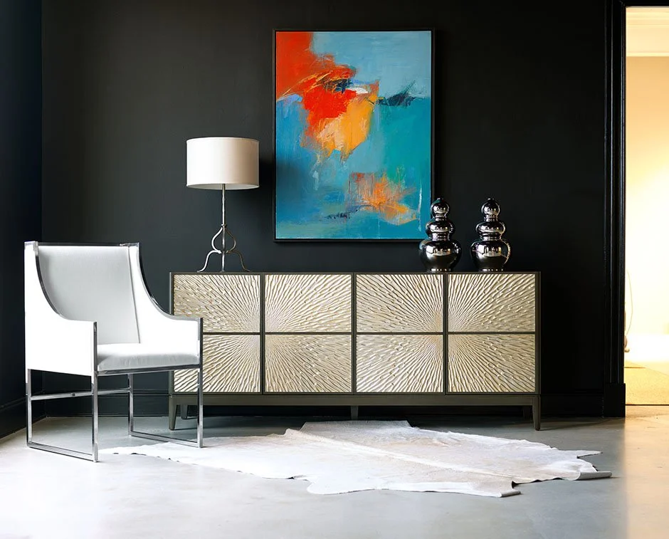 Modern interior with black wall, abstract blue and orange painting, white armchair, metallic sideboard with textured cream doors, table lamp, two black decorative vases, white rug, and doorway to a bright room.
