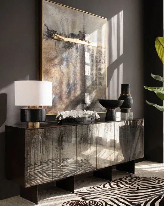 Modern living room decor with a dark-toned sideboard, abstract artwork, black and white lamps, black vases, white flowers, a zebra-patterned rug, and a large green plant, illuminated by sunlight through a window.