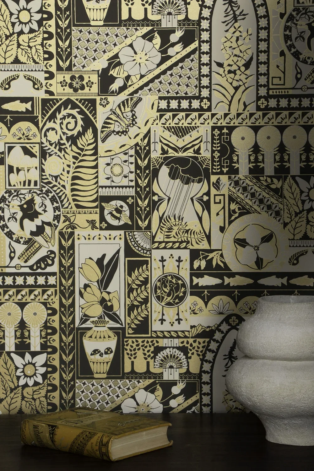 Intricate black, white, and beige patterned wallpaper with floral, bird, butterfly, mushroom, and nature motifs. A large white textured ceramic vase and a closed yellow book with a decorative cover sit on a dark wooden surface in front of the wallpaper.
