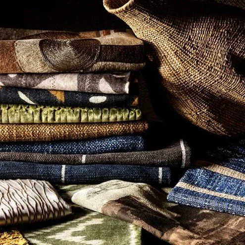A stack of folded jeans and fabric in various colors and patterns, with a brown bag on top.